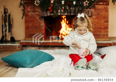 A baby girl drinks hot chocolate by the fireplace at home. A little girl on Christmas Eve or New Year's Eve at home. The concept of Christmas and New Year A baby girl drinks hot chocolate by the fireplace at home. A little girl on Christmas Eve or New Year's Eve at home. The concept of Christmas and New Year 96139854