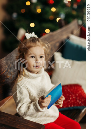 Portrait of a curly-haired little girl in a warm knitted sweater with a book in her hands against the background of a Christmas tree enjoying a cozy winter evening at home. Christmas, New Year Portrait of a curly-haired little girl in a warm knitted sweater with a book in her hands against the background of a Christmas tree enjoying a cozy winter evening at home. Christmas, New Year 96139919