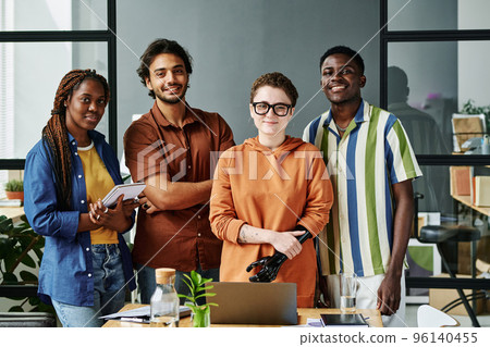 Group of young creative designers in casualwear standing by workplace with laptop and supplies in office and looking at camera Group of young creative designers in casualwear standing by workplace with laptop and supplies in office and looking at camera 96140455