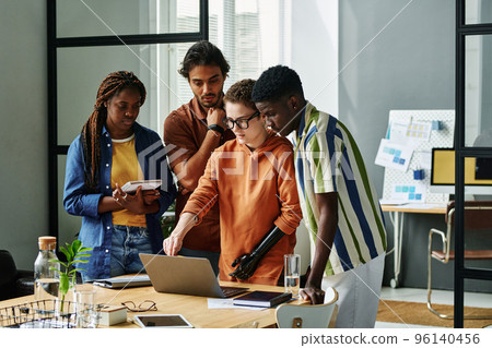 Young confident designer with partial arm pointing at laptop screen while making presentation of project plan to intercultural colleagues Young confident designer with partial arm pointing at laptop screen while making presentation of project plan to intercultural colleagues 96140456