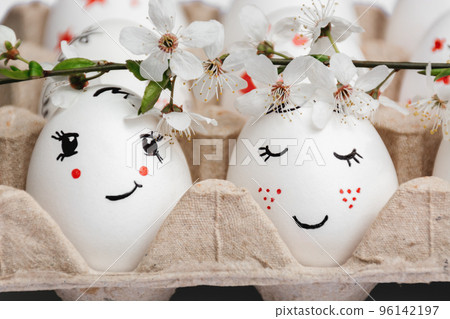 Close-up of delicately painted Easter eggs in paper eco-friendly tray. From above, sprig of spring white flowers from fruit trees. Pair of eggs looks like bride and groom. Spring style. Postcard Close-up of delicately painted Easter eggs in paper eco-friendly tray. From above, sprig of spring white flowers from fruit trees. Pair of eggs looks like bride and groom. Spring style. Postcard 96142197