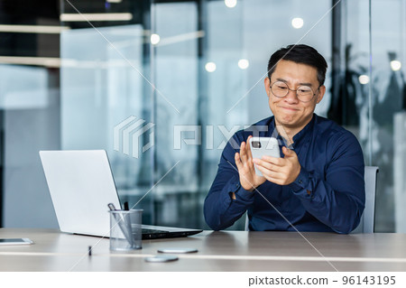 Dissatisfied asian businessman got bad news, man reading on smartphone sitting inside modern office with laptop, disappointed boss with phone upset. Dissatisfied asian businessman got bad news, man reading on smartphone sitting inside modern office with laptop, disappointed boss with phone upset. 96143195