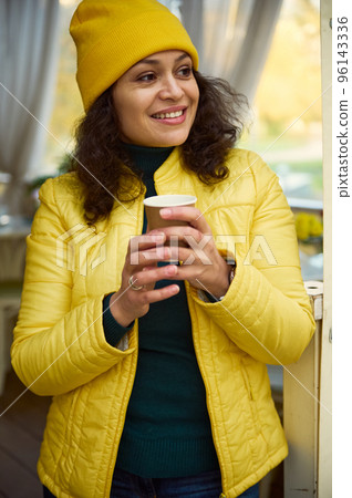 Close-up portrait of an attractive brunette multi-ethnic woman, in bright yellow hat and jacket, smiling a cheerful tothy smile looking aside, warming her hands with a takeaway hot coffee in paper cup 96143336