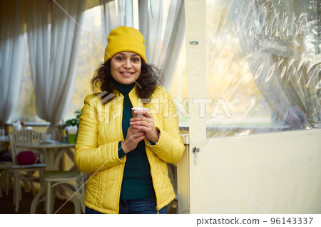 Attractive curly woman, wearing a green jacket, bright yellow hat and jacket, warming her hands with a takeaway hot aroma coffee in a disposable paper cup, smiling a beautiful toothy smile to camera 96143337