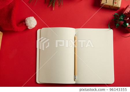 Still life with a wooden pencil on an open notepad or agenda with blank paper sheets and copy advertising space for promotional text, isolated on a red background with Christmas items and Santa's hat Still life with a wooden pencil on an open notepad or agenda with blank paper sheets and copy advertising space for promotional text, isolated on a red background with Christmas items and Santa's hat 96143355