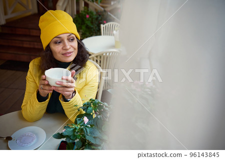 Close-up portrait of a delightful Latin American woman in yellow jacket and hat, taking a sip of hot drink, warming her hands with a cup of tea or coffee, enjoying outdoor coffee break on fall day Close-up portrait of a delightful Latin American woman in yellow jacket and hat, taking a sip of hot drink, warming her hands with a cup of tea or coffee, enjoying outdoor coffee break on fall day 96143845