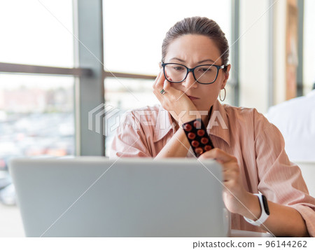 Frowning woman looks on medicine pills while working with laptop. Mental health problems, emotional burnout or headache. Modern office at co-working center. Workplace for freelancers or students. Frowning woman looks on medicine pills while working with laptop. Mental health problems, emotional burnout or headache. Modern office at co-working center. Workplace for freelancers or students. 96144262