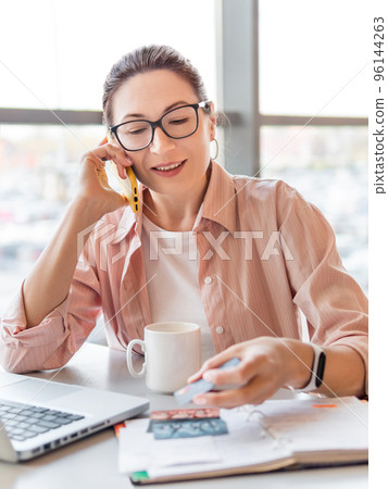 Woman works with laptop, credit cards and notepad. Smiling female is checking credit cards or ID card. Modern office with panoramic windows. Co-working center - workplace for freelancers or students. Woman works with laptop, credit cards and notepad. Smiling female is checking credit cards or ID card. Modern office with panoramic windows. Co-working center - workplace for freelancers or students. 96144263