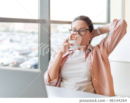 Relaxed woman drinks coffee and works with laptop. Modern office with panoramic windows at co-working center. Workplace for freelancers or students in business center. Relaxed woman drinks coffee and works with laptop. Modern office with panoramic windows at co-working center. Workplace for freelancers or students in business center. 96144264
