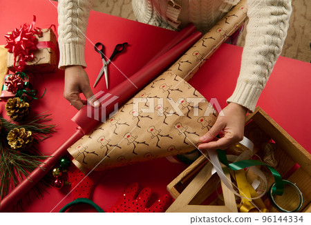 Top view woman's hands picking up gift paper, rolling out the red one and the other with deer pattern over red surface, surrounded by festive Christmas ornaments and wrapping materials. New Year. Xmas Top view woman's hands picking up gift paper, rolling out the red one and the other with deer pattern over red surface, surrounded by festive Christmas ornaments and wrapping materials. New Year. Xmas 96144334
