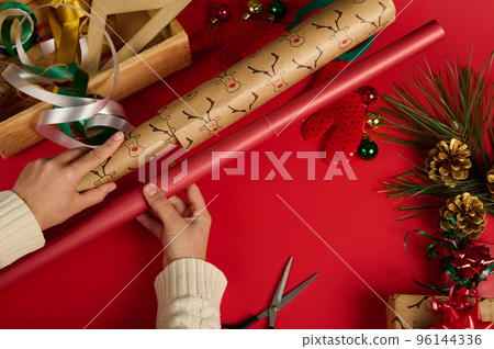Top view woman's hands hold gift paper of red color and with deer pattern, choosing wrapping paper for packing Christmas or New Year's presents, on red background. Festive decorations in wooden crate Top view woman's hands hold gift paper of red color and with deer pattern, choosing wrapping paper for packing Christmas or New Year's presents, on red background. Festive decorations in wooden crate 96144336