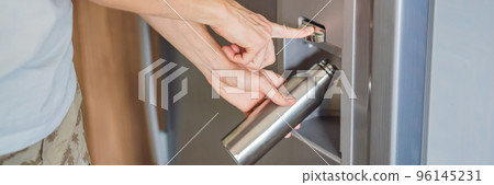 Male hand is pouring cold water and ice cubes in a metal bottle from dispenser of home fridge BANNER, LONG FORMAT 96145231
