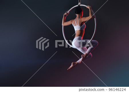 Young woman performing acrobatic element on aerial ring against dark background. Space for text 96146224