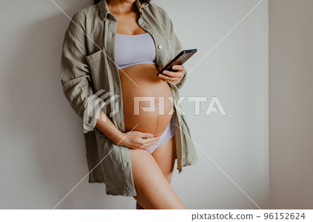 Pregnant woman using phone app for online pregnancy prenatal class or reading about motherhood wearing underwear and casual cotton shirt at home. Relaxing holding expecting belly for health. 96152624