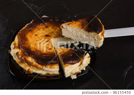 Spanish Basque cheesecake on a black plate, on a black slate, two pieces were cut off and one was raised on a knife Spanish Basque cheesecake on a black plate, on a black slate, two pieces were cut off and one was raised on a knife 96155450