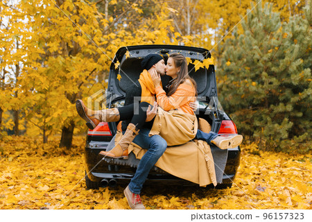 A cute young couple in love is sitting in a car with the trunk open, hugging, ready to kiss the autumn forest for the first time. The concept of travel and love A cute young couple in love is sitting in a car with the trunk open, hugging, ready to kiss the autumn forest for the first time. The concept of travel and love 96157323