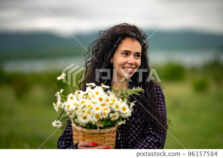 A woman stands on a green field and holds a basket with a large bouquet of daisies in her hands. In the background are mountains and a lake. 96157584