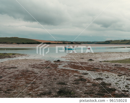 Clonakilty Bay at low tide on a day. A small blue fishing boat is anchored. Open seabed, silt and algae. Picturesque seaside landscape. 96158028