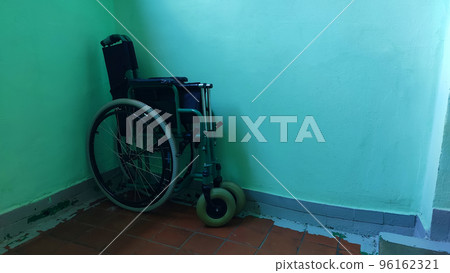 Folding wheelchair, standing folded against a green wall in the corner 96162321