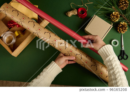 Top view woman's hands hold gift paper of red color and with deer pattern, choose wrapping paper for packing Christmas or New Year's presents over green background. Festive decorations in wooden crate Top view woman's hands hold gift paper of red color and with deer pattern, choose wrapping paper for packing Christmas or New Year's presents over green background. Festive decorations in wooden crate 96165208