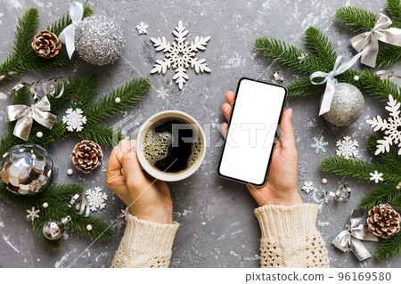 Above view of female hand holding smart phone with hot cup of coffee. Christmas decoration on colored background Above view of female hand holding smart phone with hot cup of coffee. Christmas decoration on colored background 96169580
