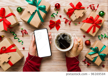 Above view of female hand holding smart phone with hot cup of coffee. Christmas decoration on colored background Above view of female hand holding smart phone with hot cup of coffee. Christmas decoration on colored background 96169584