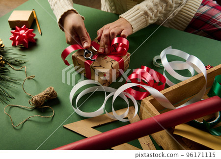 Close-up woman wraps Christmas gift box, tying up with a shiny red ribbon. Decorative tapes and bows, linen rope and ornaments on green background. New Year preparations. Packing presents. Boxing Day Close-up woman wraps Christmas gift box, tying up with a shiny red ribbon. Decorative tapes and bows, linen rope and ornaments on green background. New Year preparations. Packing presents. Boxing Day 96171115