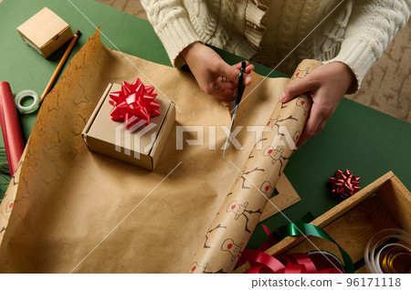 Overhead view of a woman cutting the wrapping paper while packing gifts for Christmas, New Year or any other celebration event. Decorative tapes on a wooden crate in green background. Boxing Day Overhead view of a woman cutting the wrapping paper while packing gifts for Christmas, New Year or any other celebration event. Decorative tapes on a wooden crate in green background. Boxing Day 96171118