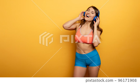 Fitness girl listens to music with headset. Joyful and happy expression on yellow background Fitness girl listens to music with headset. Joyful and happy expression on yellow background 96171908