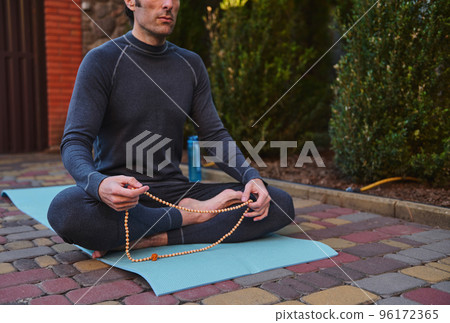 Cropped view of a yogi sitting on a fitness mat, meditating with rosary beads while practicing yoga outdoors. Body conscious. Sport. Active and healthy lifestyle. Meditation. Spiritual growth 96172365