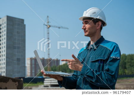 A male builder in a helmet stands against the background of a construction site with a laptop in his hands. A male builder in a helmet stands against the background of a construction site with a laptop in his hands. 96179407