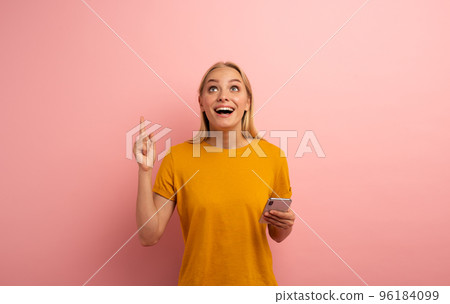 Blonde cute girl, with her smartphone, indicates something. Amazed and surprised expression face. Pink background with blank space for your text Blonde cute girl, with her smartphone, indicates something. Amazed and surprised expression face. Pink background with blank space for your text 96184099