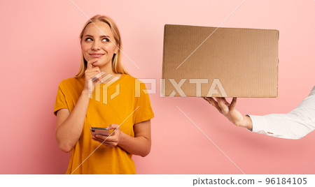 Blonde cute girl receives a box from online shop order. Surprised and pensive expression. Pink background. Blank space for your text 96184105