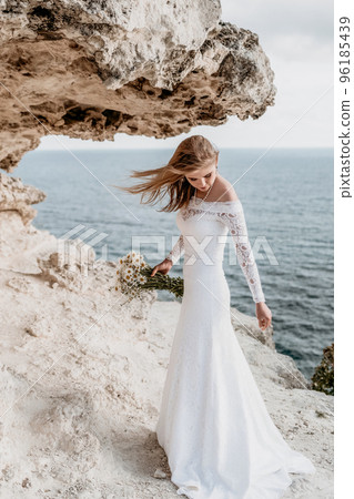 Romantic bride, blond girl in white wedding dress with open shoulders posing with sea and rocks in background. Stylish young woman standing on cliff's edge and looking into a ocean mountains at sunset 96185439