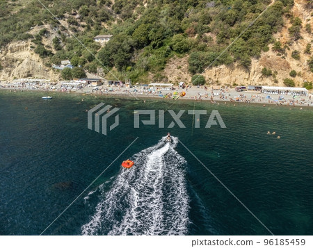 Happy people swim on air mattress behind a high speed water bike. Tourists ride on inflatable watercraft boat. Sea attraction. Water scooter skating people on large Inflatable raft. Aerial copter view 96185459