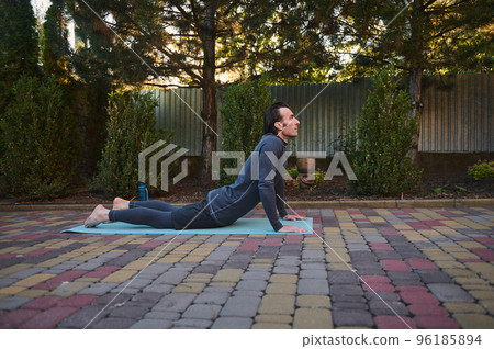 Side view a male athlete in gray activewear, a determined sportsman stretches his body on a yoga mat while practicing bodyweight training outdoors. Fitness. Stretching. Sport. Active lifestyle 96185894