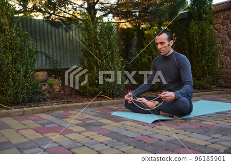 Peaceful man yogi sitting in lotus pose on a fitness mat, meditating with rosary beads outdoors at sunset. Yoga practice. Meditation. Enlightenment. Body conscious. Active and healthy lifestyle Peaceful man yogi sitting in lotus pose on a fitness mat, meditating with rosary beads outdoors at sunset. Yoga practice. Meditation. Enlightenment. Body conscious. Active and healthy lifestyle 96185901