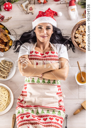 Satisfied dark-haired female cook in a Christmas apron and Santa hat,lying on the floor, surrounded by gingerbread, Linz cakes, on the other side 96194536