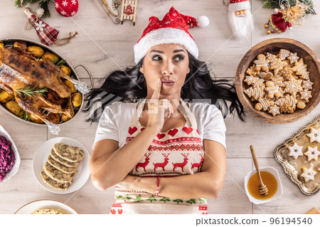Pensive female cook in a Christmas apron and Santa hat,lying on the floor, surrounded by gingerbread, Linz cakes, on the other side 96194540