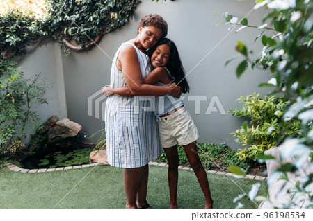 Granny and granddaughter embracing each other while standing in backyard Granny and granddaughter embracing each other while standing in backyard 96198534