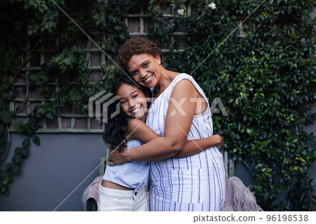 Granny and grandchild embrace and look at the camera. Grandma and granddaughter hugging and smiling while standing together outside.	 96198538