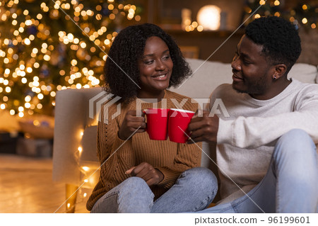 Glad young african american couple cheers with cups of hot drink, enjoy holiday and free time, sit on floor Glad young african american couple cheers with cups of hot drink, enjoy holiday and free time, sit on floor 96199601