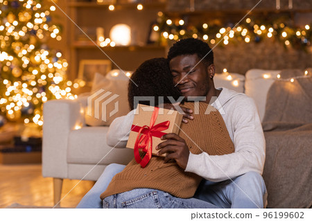 Glad young african american guy hugs wife and hold box with gift near Christmas tree with toys and luminous garlands 96199602