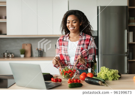 Glad black young lady in casual prepares salad from organic vegetables and watch video lesson of recipe 96199673
