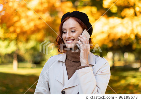 Cheerful pretty caucasian millennial female with red hair in raincoat and hat calling by phone, enjoy autumn 96199678