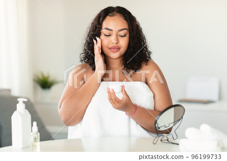 Facial skin care concept. Young black overweight woman applying moisturizer holding cream jar, sitting in bedroom 96199733