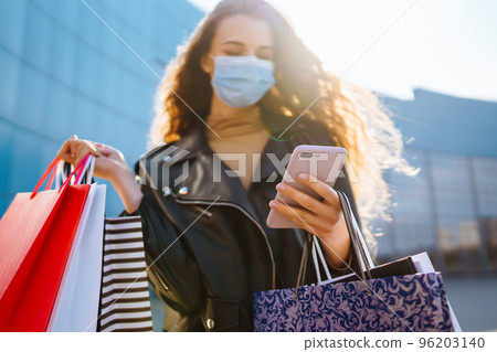 Smiling woman in protective medical mask with shopping bags using her phone. Young woman after. Smiling woman in protective medical mask with shopping bags using her phone. Young woman after. 96203140