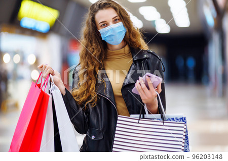 Young woman in protective mask using mobile phone while holding shopping bag in department store. 96203148
