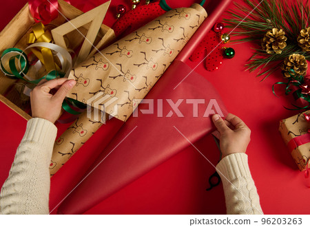 View from above of woman's hands, rolling out a red gift paper and with deer pattern, wrapping, packing Christmas and New Year presents. Boxing Day. Packaging. Christmastime. Diy presents 96203263