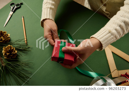 Close-up woman tying up present, wrapped in red paper with green decorative ribbon. Packing Christmas and New Year gifts. Happy holiday present, surprise. Magic time. Celebration winter event Close-up woman tying up present, wrapped in red paper with green decorative ribbon. Packing Christmas and New Year gifts. Happy holiday present, surprise. Magic time. Celebration winter event 96203273